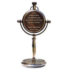 Puede incluir: Un reloj de escritorio decorativo de color lat&oacute;n, con una esfera redonda y un soporte. La esfera del reloj tiene la inscripci&oacute;n: "Never underestimate the difference you have made and the lives you have touched. Happy Retirement Ronny Paul."