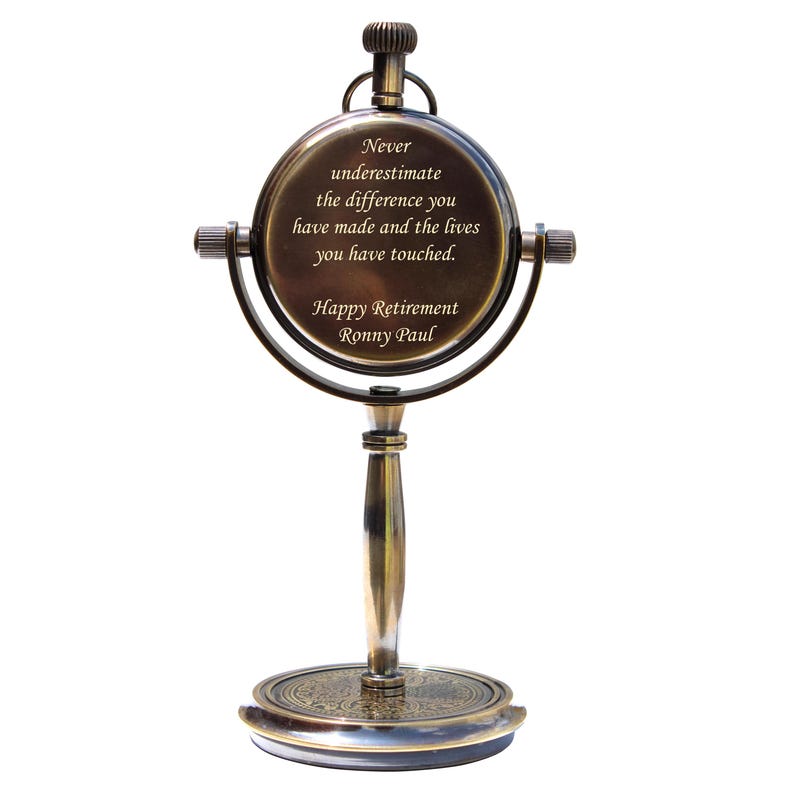 Puede incluir: Un reloj de escritorio decorativo de color lat&oacute;n, con una esfera redonda y un soporte. La esfera del reloj tiene la inscripci&oacute;n: "Never underestimate the difference you have made and the lives you have touched. Happy Retirement Ronny Paul."