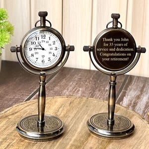 Puede incluir: Reloj de escritorio de estilo antiguo con esfera redonda y acabado de bronce detallado. El reloj tiene una esfera blanca con n&uacute;meros y manecillas negras. La parte posterior del reloj tiene una inscripci&oacute;n que dice: "Thank you John for 35 years of service and dedication. Congratulations on your retirement!"