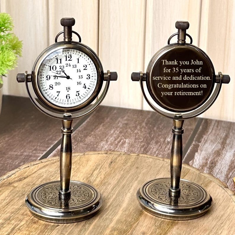 Puede incluir: Reloj de escritorio de estilo antiguo con esfera redonda y acabado de bronce detallado. El reloj tiene una esfera blanca con n&uacute;meros y manecillas negras. La parte posterior del reloj tiene una inscripci&oacute;n que dice: "Thank you John for 35 years of service and dedication. Congratulations on your retirement!"