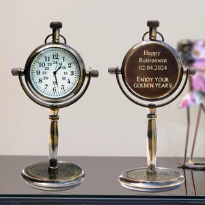 Puede incluir: Un reloj de escritorio de estilo vintage con esfera redonda y base decorativa. La esfera muestra la hora, mientras que la parte trasera del reloj tiene el texto "Happy Retirement 02.04.2024 ENJOY YOUR GOLDEN YEARS!" en tono dorado.