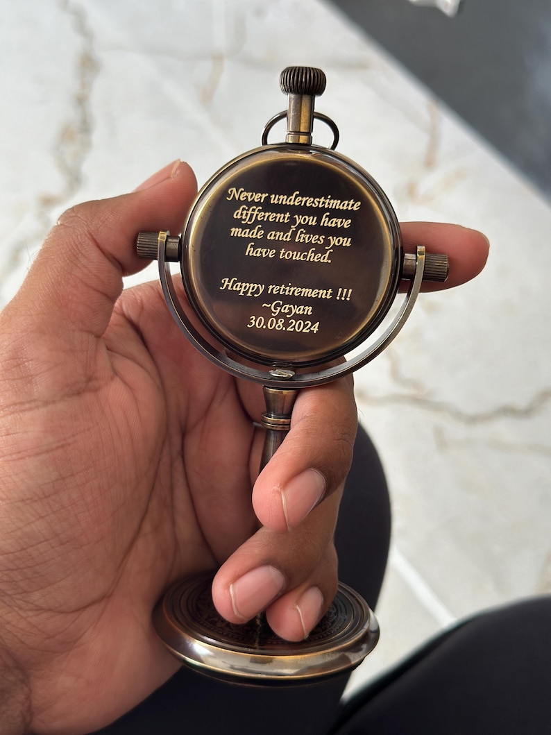 Puede incluir: Un reloj de escritorio de lat&oacute;n de estilo vintage con dise&ntilde;o de br&uacute;jula. La esfera del reloj presenta el texto grabado: "Never underestimate different you have made and lives you have touched. Happy retirement !!! ~Gayan 30.08.2024."
