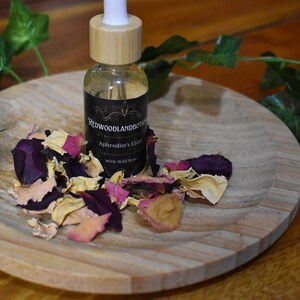 May include: A small glass bottle with a dropper, filled with a clear liquid, sits on a wooden plate with dried rose petals. The bottle label reads "Redwoodland Botanicals Aphrodite's Elixir with Wild Rose".
