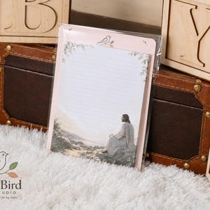 May include: A rectangular notepad in a clear plastic sleeve featuring a light pink border, a small bird illustration at the top, and a printed scene at the bottom showing a figure in a robe sitting on a rock overlooking a landscape with lined paper.