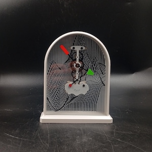 May include: A white framed kinetic art piece with a black and white wave pattern. The piece features a central mechanical element with red, green, and black accents. The word "Regale" is visible on a white tag.