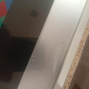May include: A close-up shot of a white, rectangular frame with a light brown, textured edge. The frame is positioned diagonally, with a dark, reflective surface visible on the left side. A colorful geometric pattern is partially visible.