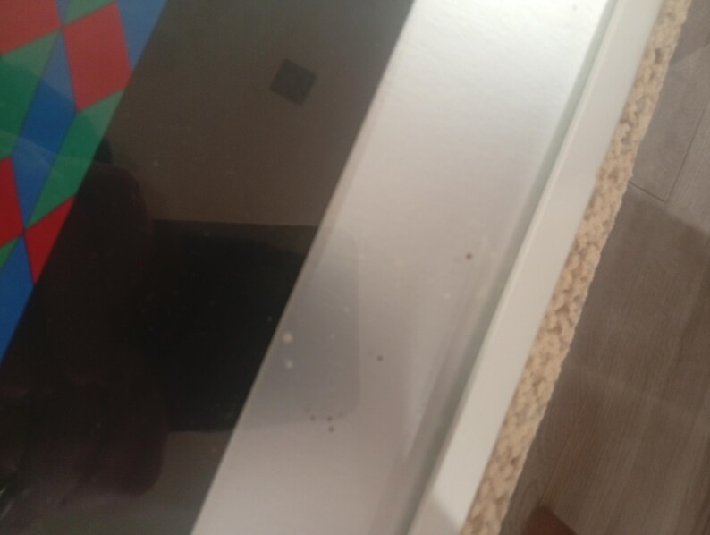 May include: A close-up shot of a white, rectangular frame with a light brown, textured edge. The frame is positioned diagonally, with a dark, reflective surface visible on the left side. A colorful geometric pattern is partially visible.