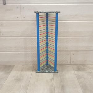 May include: A tall, rectangular sculpture with a blue frame and colorful, angled lines. The base and top are a speckled gray, and the lines are red, yellow, and green. The sculpture stands on a light wood floor.