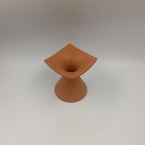 Puede incluir: Un jarrón de cerámica hecho a mano de color terracota. El jarrón tiene una parte superior cuadrada ensanchada y una base de pedestal redondeada. La abertura tiene forma de embudo. El jarrón es de un tono cálido y terroso.