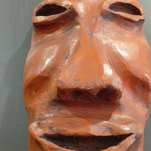 Pode incluir: Uma m&aacute;scara de terracota com um largo sorriso cheio de dentes. A m&aacute;scara tem um nariz grande e proeminente e olhos fundos.