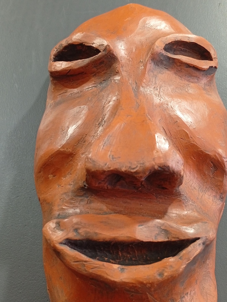 Pode incluir: Uma m&aacute;scara de terracota com um largo sorriso cheio de dentes. A m&aacute;scara tem um nariz grande e proeminente e olhos fundos.