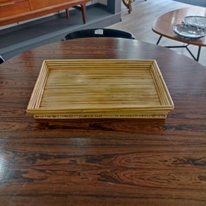 May include: A rectangular tray made of woven natural bamboo sticks. The tray is light brown in color and has a smooth, polished finish. The tray is sitting on a dark brown wooden table.