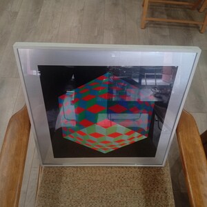 May include: A framed artwork featuring a geometric design of a cube with red, green, and blue diamond patterns. The artwork is set against a black background and is encased in a silver frame. The artwork is resting on a chair.