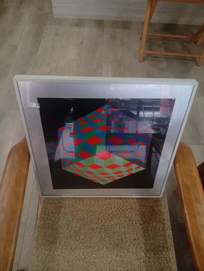 May include: A framed artwork featuring a geometric design of a cube with red, green, and blue diamond patterns. The artwork is set against a black background and is encased in a silver frame. The artwork is resting on a chair.