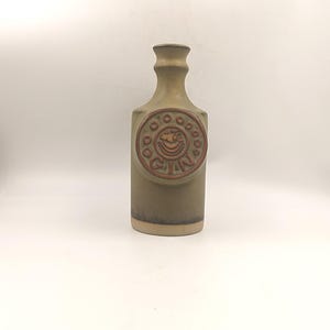 Könnte beinhalten: Eine Keramik-Gin-Flasche in Olivgrün mit rechteckigem Körper und schmalem Hals. Ein kreisförmiges Emblem in Rotbraun trägt die Aufschrift "GIN". Die Flasche hat eine Vintage-Ästhetik.