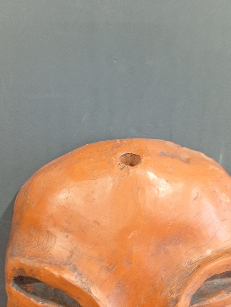 Pode incluir: Uma m&aacute;scara de terracota com um furo no topo para pendurar. A m&aacute;scara tem uma superf&iacute;cie arredondada e lisa e apresenta dois olhos grandes esculpidos.
