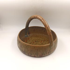 Op de afbeelding: Een kleine, ronde keramische kom met een handvat. De kom is bruin met rode en oranje vlekken en een gevlekt bruin handvat.