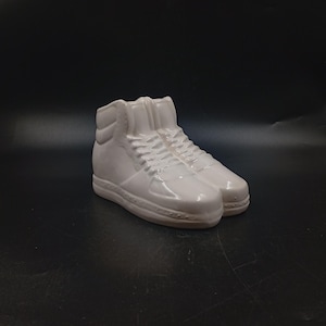 Peut inclure: Une paire de baskets montantes blanches en céramique. Les chaussures ont une finition brillante, des lacets et une semelle texturée. Les baskets sont un objet décoratif, peut-être une figurine ou une petite sculpture, sur fond sombre.