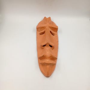 Peut inclure: Un masque de couleur terre cuite avec un visage stylisé. Le masque présente un nez proéminent, une moustache et une bouche tombante, avec des orbites sculptées et un menton pointu. Le masque est probablement fait à la main et a une finition lisse et mate.