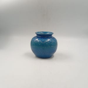 May include: A round, blue ceramic vase with a textured surface and a wide opening. The vase features a detailed pattern around the middle and top, adding visual interest. The color is a vibrant teal, suitable for home decor.