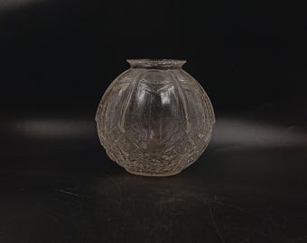Op de afbeelding: Een heldere, ronde glazen vaas met een getextureerd, patroon oppervlak. De vaas heeft een brede, bolvormige body en een smalle, gedetailleerde rand. Het ontwerp bevat herhalende geometrische en bloemmotieven, waardoor een vintage esthetiek ontstaat.
