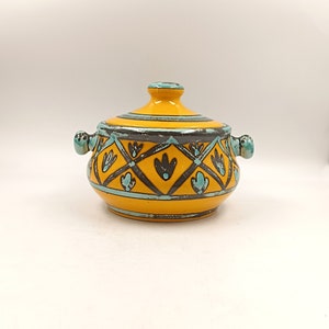 Pode incluir: Um pote de cerâmica amarelo com tampa, com um padrão geométrico azul e preto. O pote tem duas alças nas laterais.