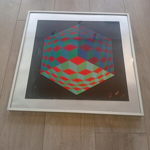 May include: Framed geometric art print featuring a 3D cube design. The artwork showcases a vibrant pattern of red, green, and blue diamond shapes against a black background. The print is encased in a white frame, suitable for wall decor.