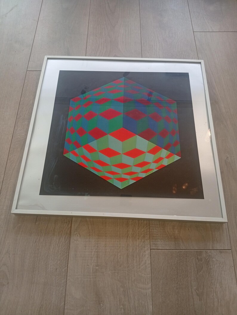 May include: Framed geometric art print featuring a 3D cube design. The artwork showcases a vibrant pattern of red, green, and blue diamond shapes against a black background. The print is encased in a white frame, suitable for wall decor.