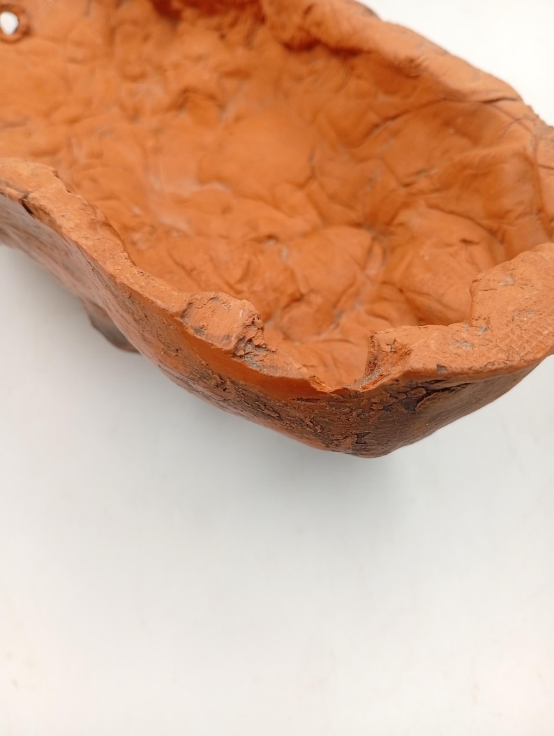 Pode incluir: Um vaso de terracota com uma superf&iacute;cie &aacute;spera e texturizada. O vaso tem formato oval e uma abertura larga e rasa.
