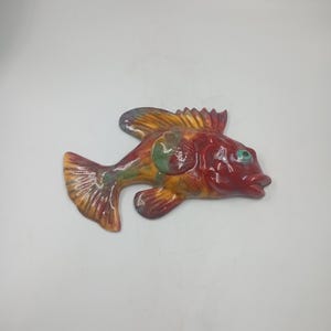 Könnte beinhalten: Eine Keramik-Fischskulptur mit glänzendem Finish. Der Fisch hat einen roten Körper, gelbe und orangefarbene Flossen und einen grünen Fleck. Er hat ein kleines, blaues Auge und ist vor einem weißen Hintergrund platziert. Dieses dekorative Stück eignet sich für die Inneneinrichtung.