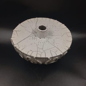 May include: A handcrafted, off-white ceramic vessel with a circular top and a cylindrical opening. The surface features a crackle glaze pattern and a textured, scalloped edge. The vessel is likely used for decorative purposes.