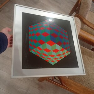 May include: Framed geometric art print featuring a 3D cube design. The cube is composed of red, green, and blue diamond shapes against a black background. The artwork is encased in a white frame.