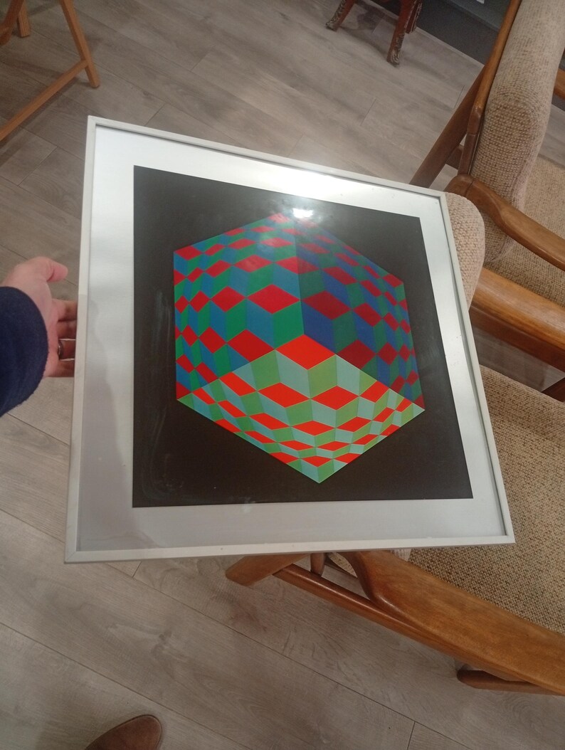 May include: Framed geometric art print featuring a 3D cube design. The cube is composed of red, green, and blue diamond shapes against a black background. The artwork is encased in a white frame.