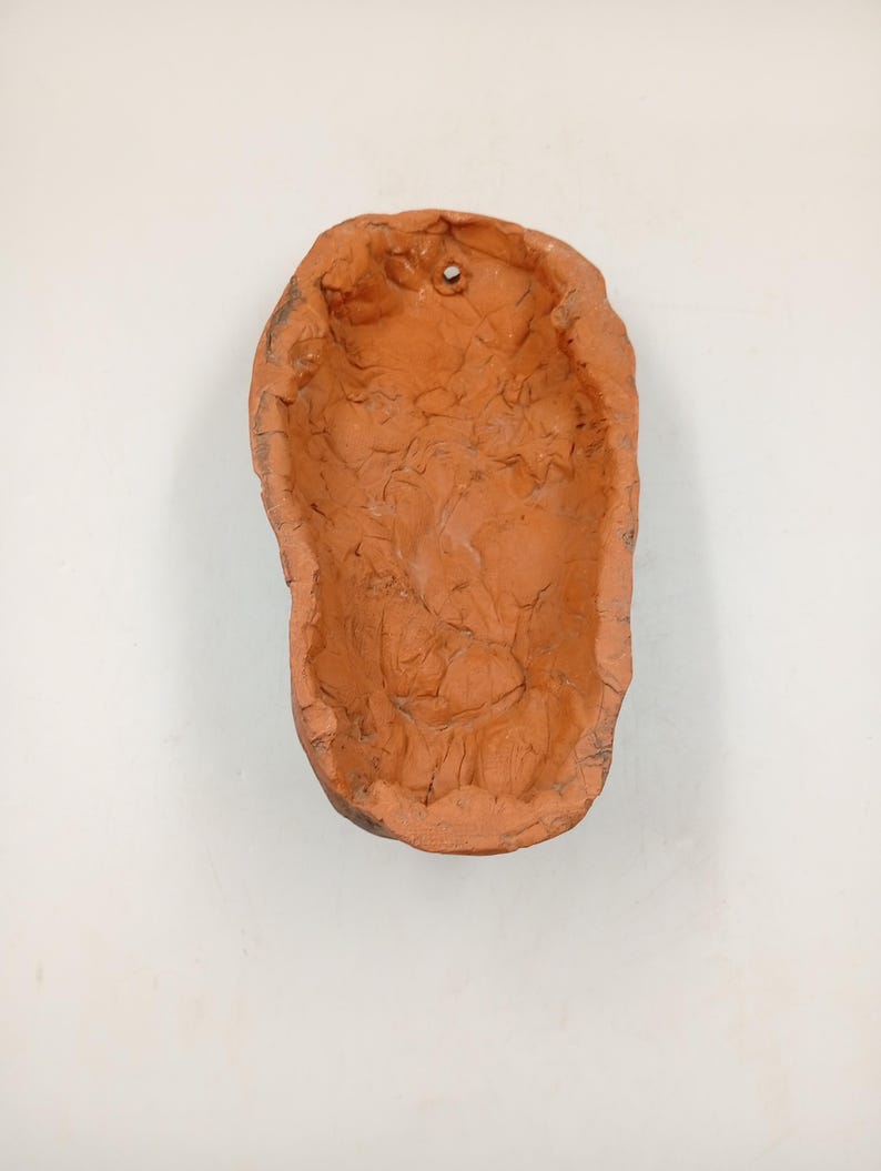 Pode incluir: Um vaso de argila de cor terracota com uma superf&iacute;cie &aacute;spera e texturizada. O vaso tem um furo na parte superior para pendurar.