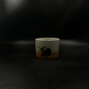 May include: A small, cylindrical ceramic cup with a cream-colored exterior. The cup features a dark, abstract design with brown and black accents. The bottom portion has a textured, yellow-brown band. The cup is suitable for beverages.