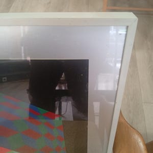 May include: A framed artwork with a white wooden frame. The artwork features a geometric design with red, green, and blue diamond shapes. The frame is rectangular and has a clean, modern aesthetic.