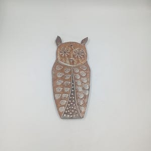 May include: A brown, owl-shaped wall hanging with a textured surface. The owl has large, round eyes, small ear-like protrusions, and a patterned design on its body. The design includes leaf-like shapes and a dotted pattern.
