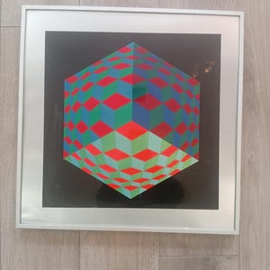 May include: Framed geometric art print featuring a 3D cube design. The cube is composed of red, green, and blue diamond shapes against a black background. The artwork is encased in a silver frame, creating a modern aesthetic.