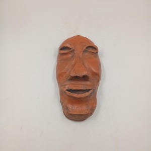 Peut inclure: Un masque en terre cuite avec un visage sculpté, les yeux fermés et une bouche légèrement ouverte. Le masque est de couleur brun rougeâtre et a une texture rugueuse.
