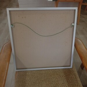 May include: Back of a framed artwork with a silver metal frame. The artwork's backing is a light brown color, with a green wire for hanging. The frame is resting on a woven chair.