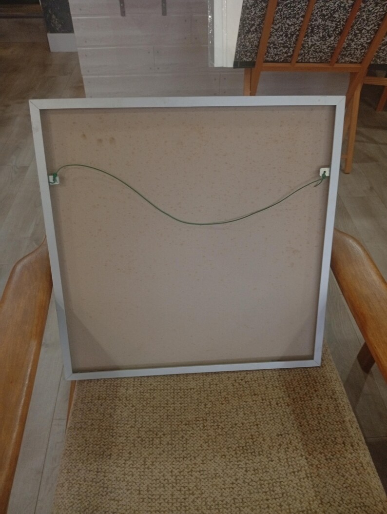 May include: Back of a framed artwork with a silver metal frame. The artwork's backing is a light brown color, with a green wire for hanging. The frame is resting on a woven chair.