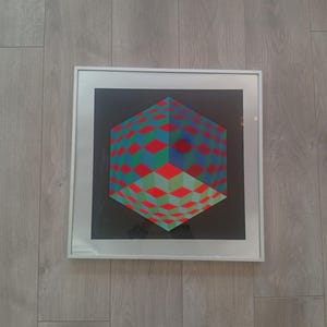 May include: Framed geometric art print featuring a three-dimensional cube design. The cube is composed of red, green, and blue diamond shapes against a black background. The artwork is encased in a white frame.