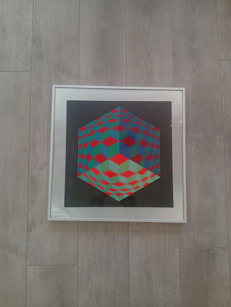 May include: Framed geometric art print featuring a three-dimensional cube design. The cube is composed of red, green, and blue diamond shapes against a black background. The artwork is encased in a white frame.