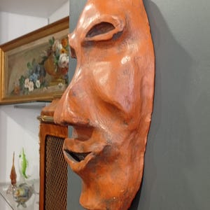 Pode incluir: Uma m&aacute;scara de terracota com um rosto humano estilizado. A m&aacute;scara &eacute; pintada em tons de laranja e marrom, e tem um nariz grande e proeminente.