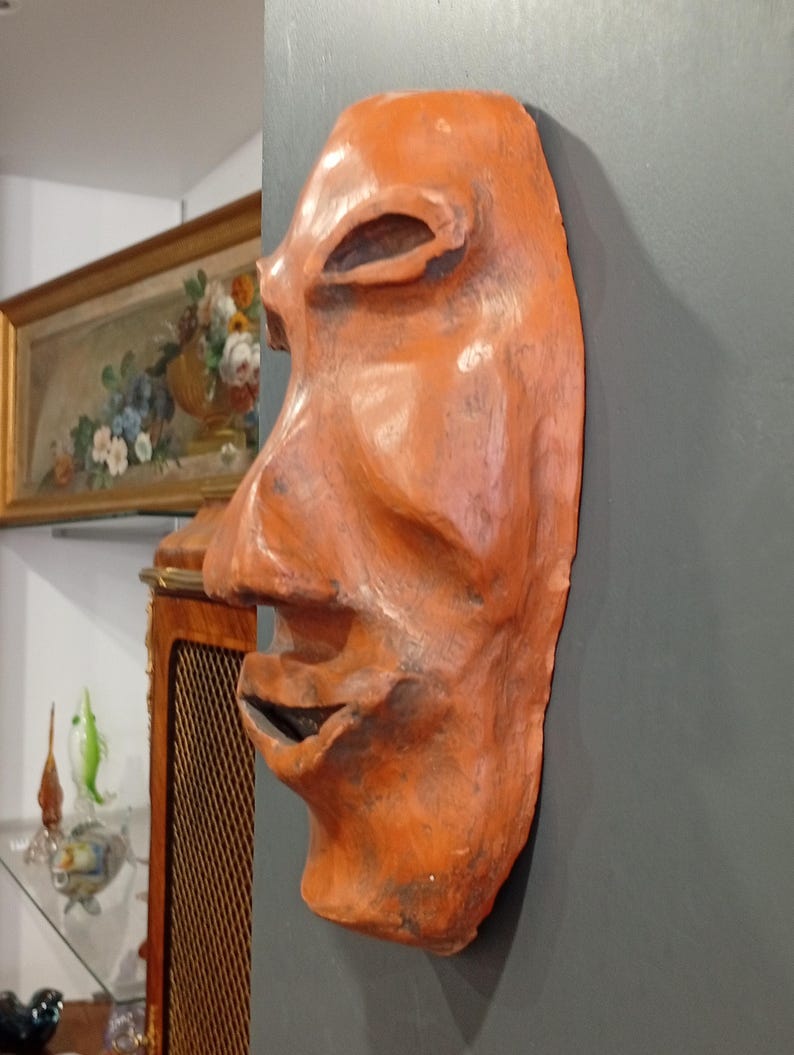 Pode incluir: Uma m&aacute;scara de terracota com um rosto humano estilizado. A m&aacute;scara &eacute; pintada em tons de laranja e marrom, e tem um nariz grande e proeminente.