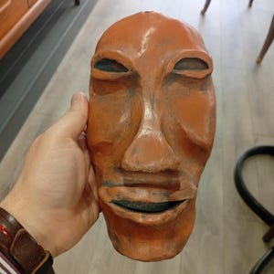 Pode incluir: Uma m&aacute;scara de madeira esculpida &agrave; m&atilde;o com um rosto humano. A m&aacute;scara &eacute; pintada em tons de laranja e marrom. Os olhos s&atilde;o ocos e a boca est&aacute; ligeiramente aberta.