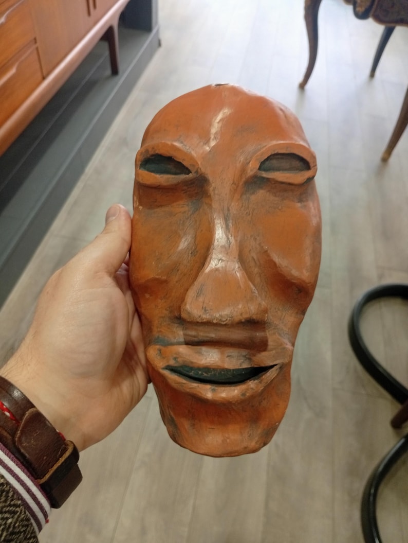 Pode incluir: Uma m&aacute;scara de madeira esculpida &agrave; m&atilde;o com um rosto humano. A m&aacute;scara &eacute; pintada em tons de laranja e marrom. Os olhos s&atilde;o ocos e a boca est&aacute; ligeiramente aberta.