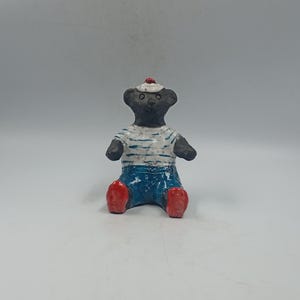 May include: A ceramic bear figurine in a seated position, wearing a sailor outfit. The bear has a black body, a white and blue striped shirt, blue trousers, red shoes, and a white hat with a red accent.