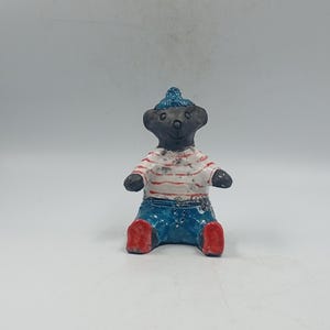 May include: A small, handcrafted bear figurine. The bear is wearing a blue knitted hat, a white shirt with red stripes, blue jeans, and red shoes. The bear is sitting with its legs outstretched.