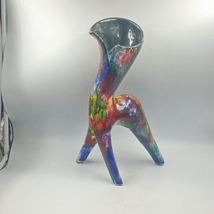 Puede incluir: Un jarrón de cerámica artesanal único con un diseño abstracto distintivo. El jarrón presenta un cuello alto y curvo y tres patas. La superficie está pintada con una vibrante mezcla de colores, incluyendo rojo, azul, verde y amarillo, con un interior gris oscuro.